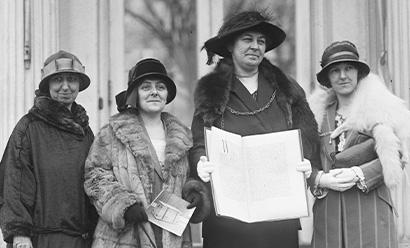 Black and white photograph of four white women dressed in fine traveling dresses, coats and hats posing outside with one woman holding open a large folio-like object with writing on the pages.