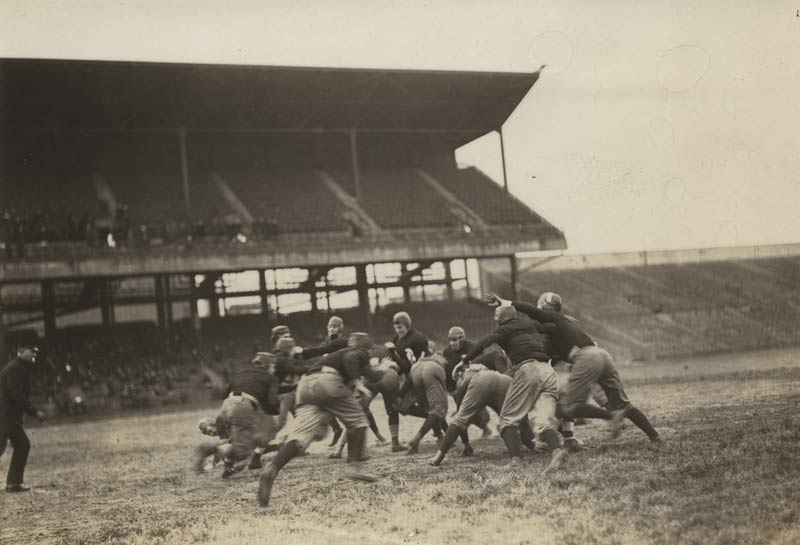Black and white action shot of a group of North American football players mid-play, with spectators in the stands behind them.