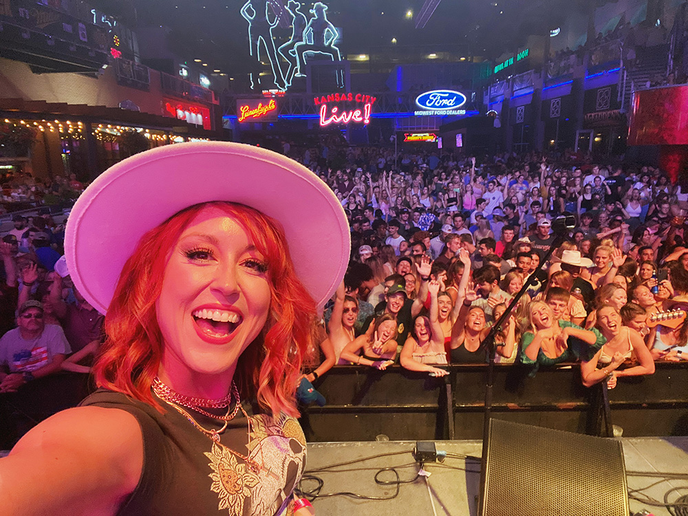 Selfie photo of a white woman with red hair wearing a pink wide-brimmed hat. Behind her is a crowd of people crammed into a theater.