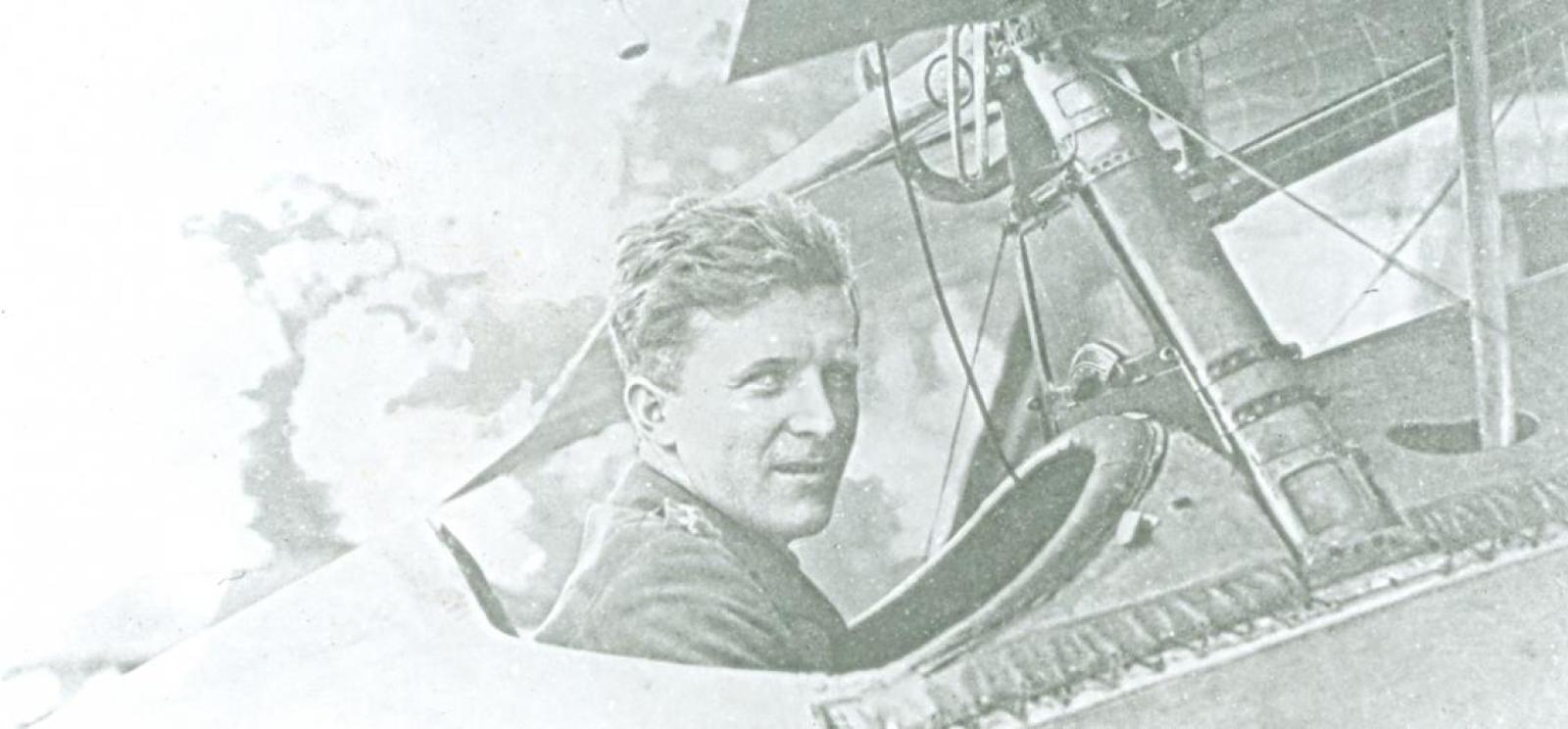 Black and white photograph of a WWI-era fighter pilot in the cockpit of a plane, looking at the viewer.