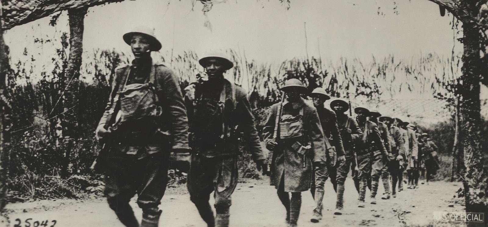 Black and white photograph of a line of Black American WWI soldiers walking through the woods
