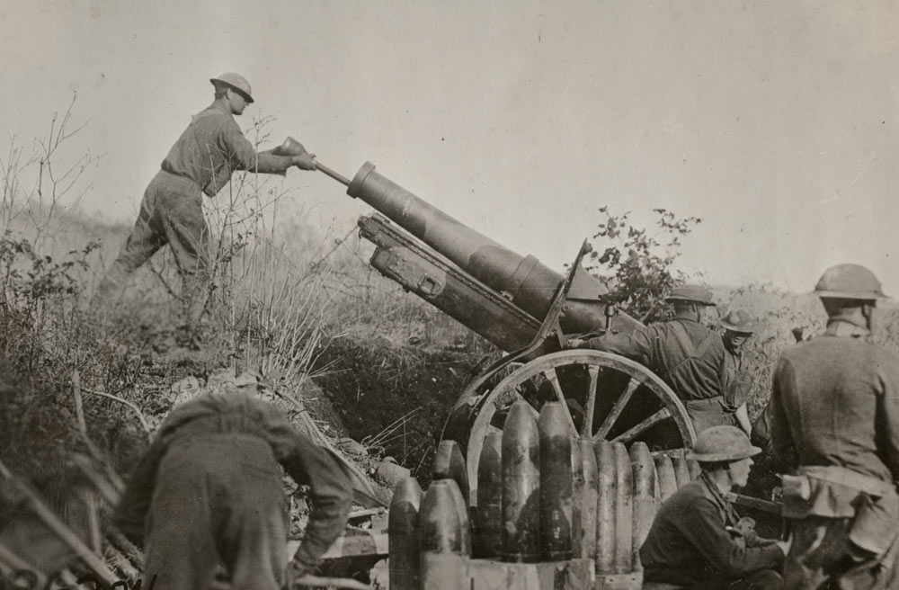 Soldiers surrounding a long, thin howitzer gun in a grassy dip. One soldier is loading ammunition into the business end of the gun.