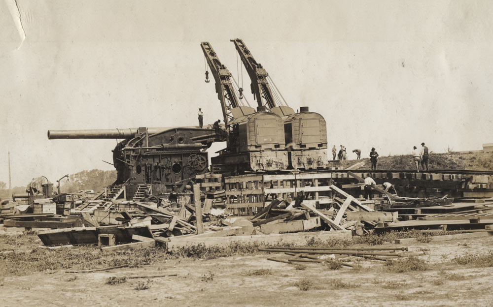 Photographie sephia d'un chantier de construction. Deux grues et des échafaudages entourent un canon de la taille d'une maison.