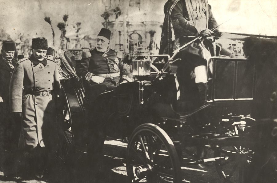 Black and white photograph of a gleaming horse carriage carrying an old portly bearded man wearing a fez and a decorated military uniform.