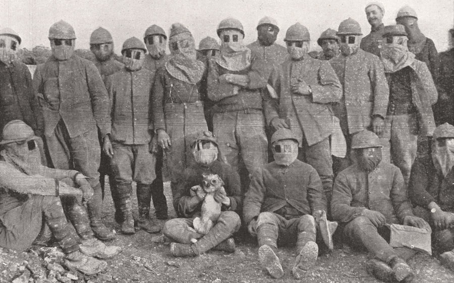 A large group of soldiers wearing early model cloth gas masks posing for a photograph. Several of the soldiers are seated in front. One of the seated soldiers holds a small dog in his lap, which is also wearing a gas mask.