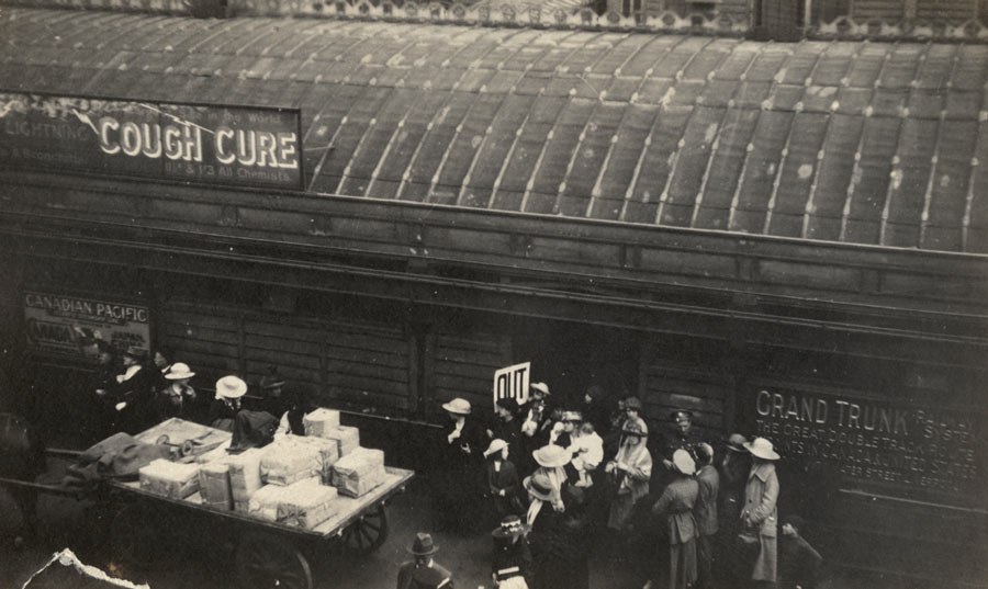 Black and white photo of a crowd standing on a railway platform surrounded by signs and advertisements