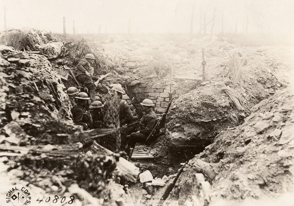 Faded black and white photograph of the inside of a narrow trench. A handful of soldiers wearing steel helmets sit or stand inside.