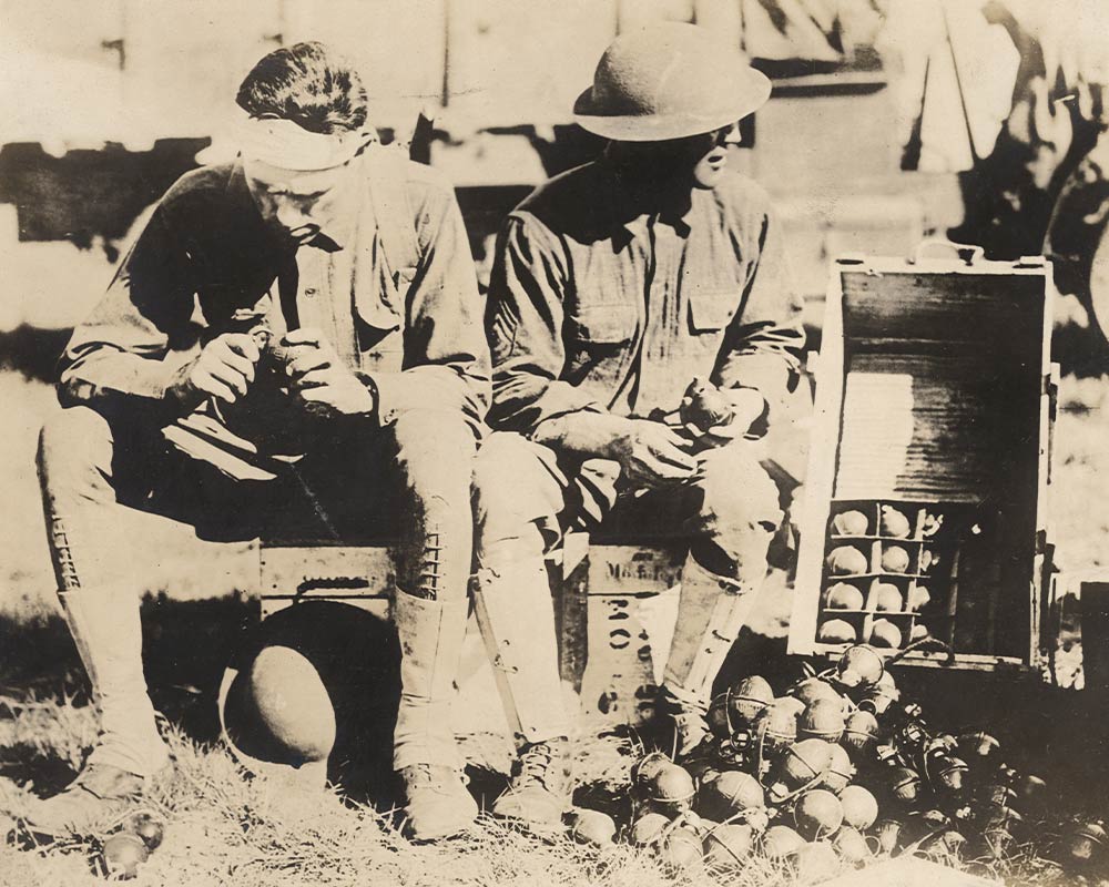 Two soldiers seated next to a large box of hand grenades, putting fuses onto the grenades.