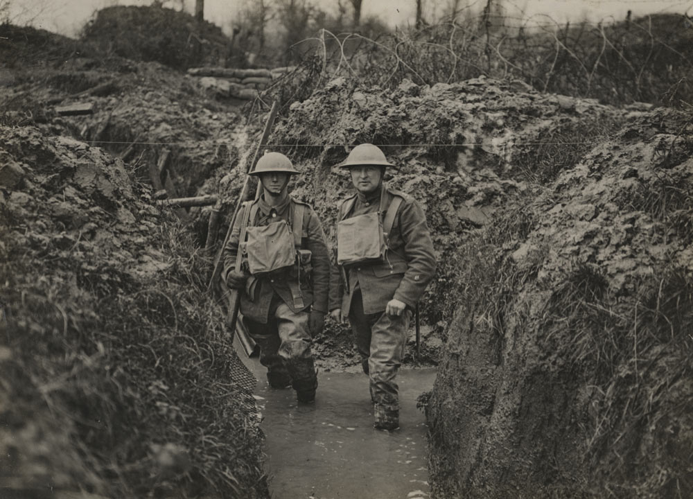 Black and white photograph of two men in military combat gear and steel helmets walking toward the viewer in a trench. They are up to their ankles in water.