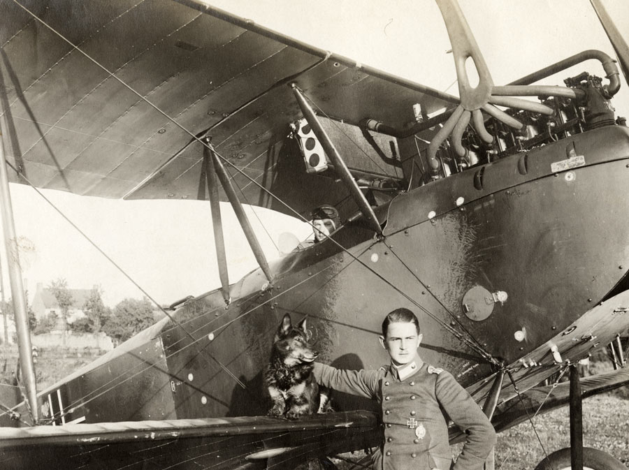 Photographie en noir et blanc d'un avion de la Première Guerre mondiale avec un pilote dans le siège. Un autre soldat se tient sur le sol devant l'aile, caressant un chien qui est assis sur l'aile.