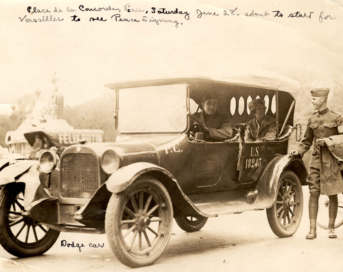 Sepia photograph of a WWI-era automobile being driven by a white man in military uniform with a white woman sitting in the back seat. Another white man in military uniform walks alongside chatting with the woman.