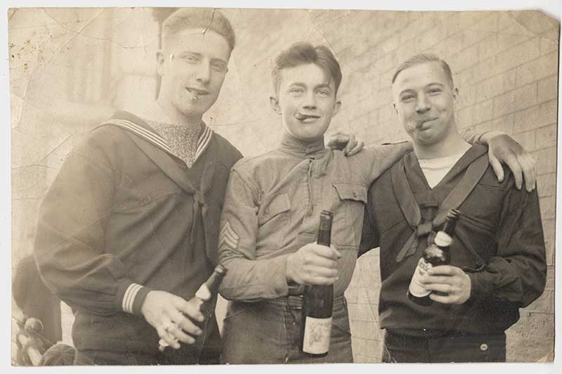 Black and white photograph of two US sailors and one US soldiers. All three hold beer bottles in their hands and cigars in their mouths as they smile at the camera.