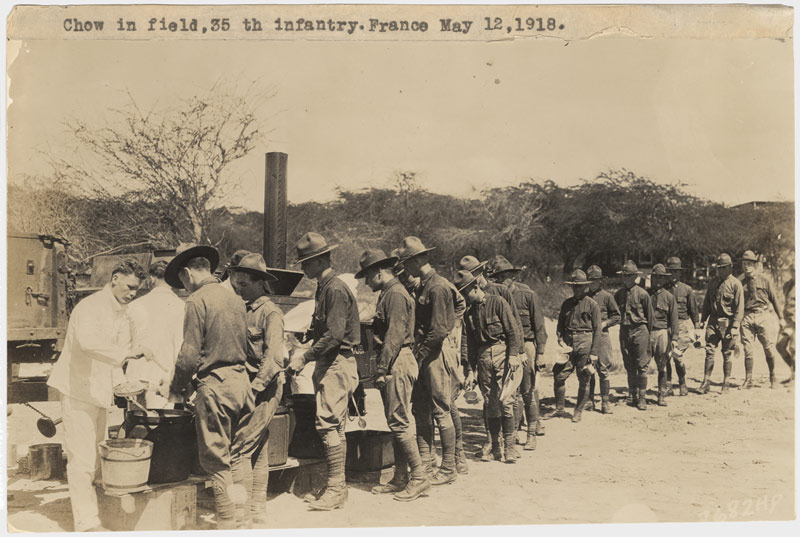 Sepia photo of several soldiers serving food to a long line of soldiers in an open field.
