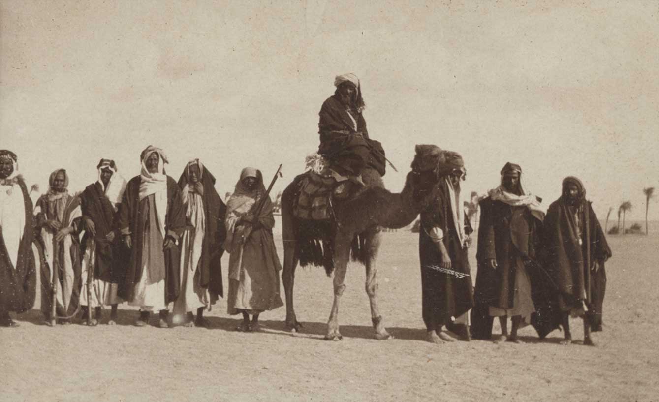 Black and white photograph of Arab men standing together with another man riding a camel at center