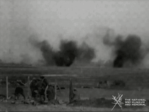 Animated gif of black and white film footage. Smoke dissipates from a bomb or cannon shot in the background. A man in military uniform walks into frame and then sheepishly walks back out.