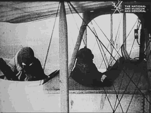 Animated gif of black and white film footage. Two men sitting in an open biplane. One is piloting. The other one stands up and throws a bomb over the side of the plane.