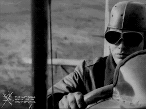 Animated gif of black and white film footage. Closeup on a man in aviator hat and goggles piloting a plane taking off.