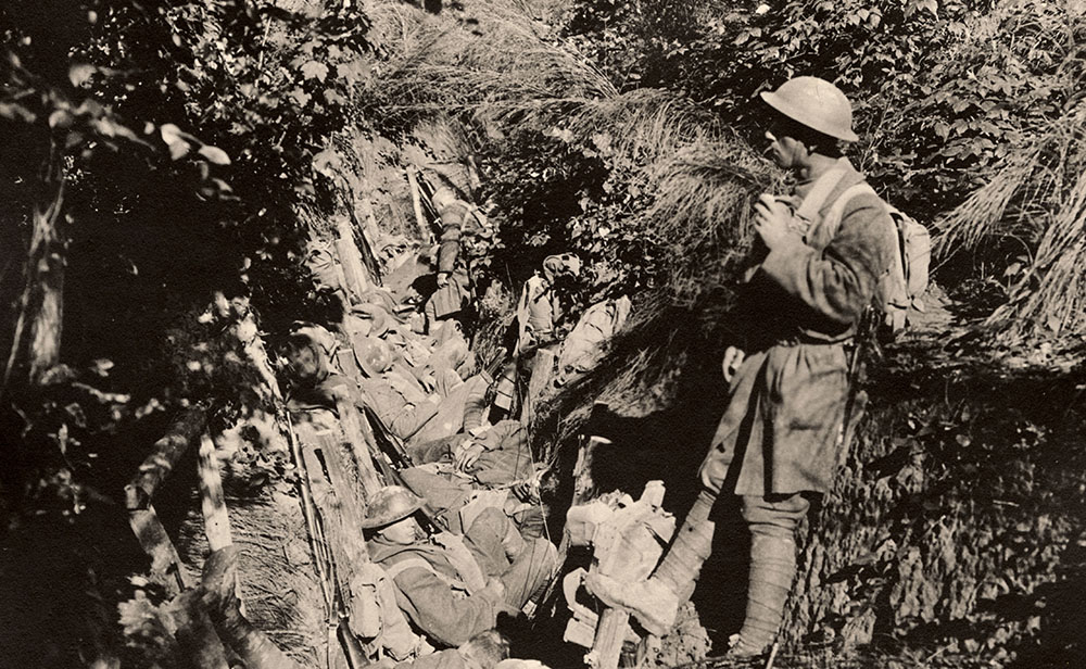 Black and white photograph of men in WWI-era military combat gear sitting or standing in a trench.