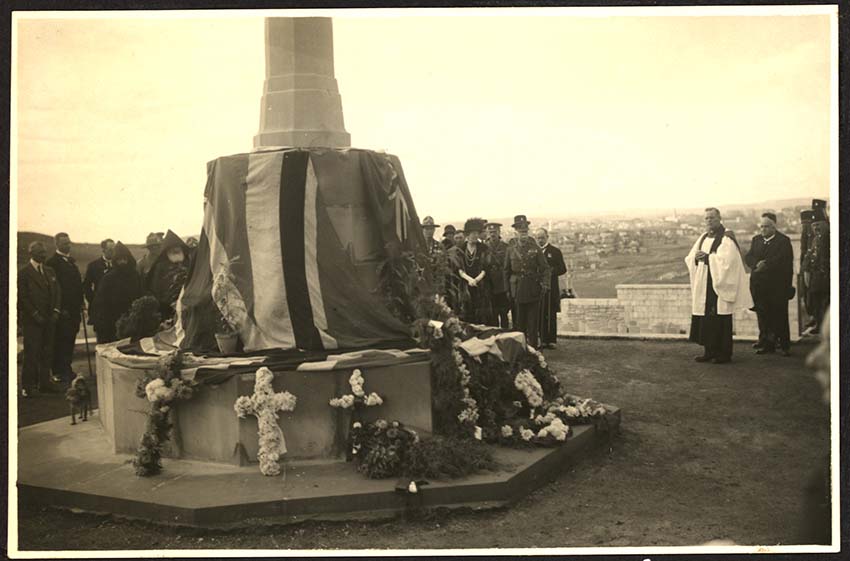 Photographie sépia d'un grand monument en pierre drapé de drapeaux et de fleurs. Un chef religieux se tient près d'elle devant une foule de personnes en deuil.