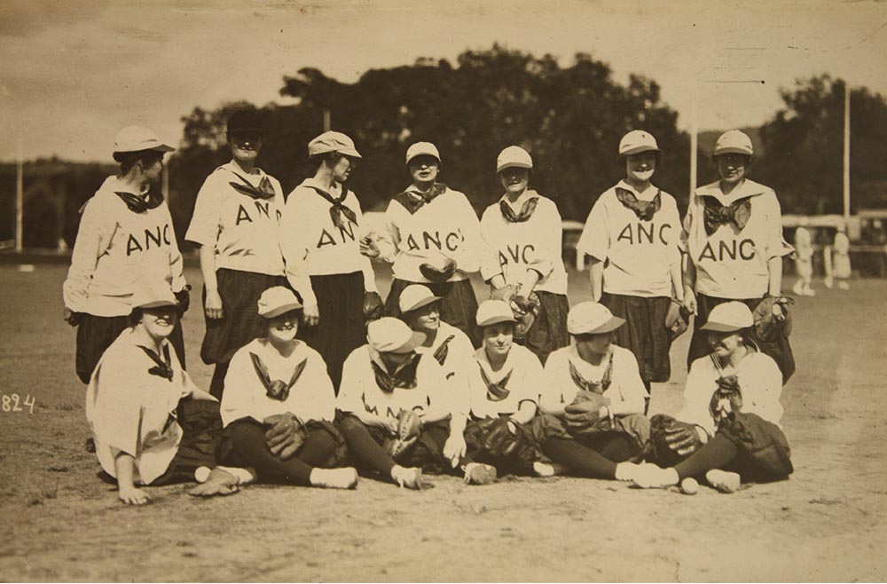 Sepia photograph of a group of white women lined up for a team photo. They are wearing loose baseball shirts and trousers and baseball caps.