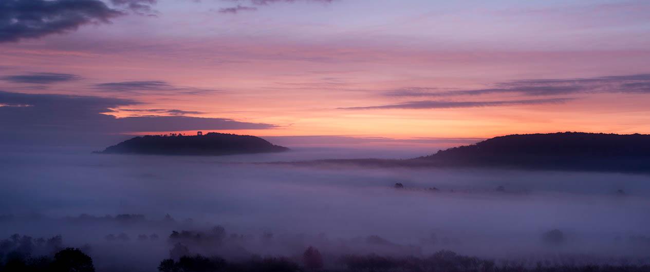 Modern photograph of an empty field and wood at dawn. The ground is covered by a thick mist.