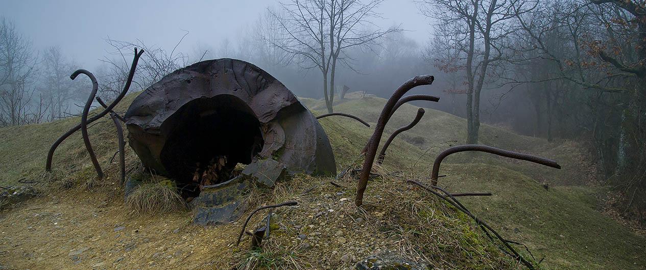Modern photograph of the remains of a concrete structure and steel rebar sticking up from the ground in an empty, misty field surrounded by trees.