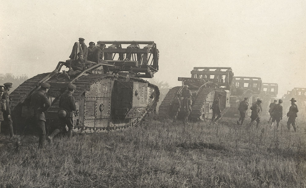Black and white photograph of a line of small tanks going through a field. Soldiers ride on the tanks and also walk alongside.