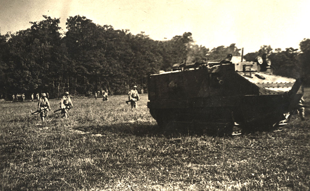 Black and white photograph of a WWI-era tank in a field followed by soldiers on foot.