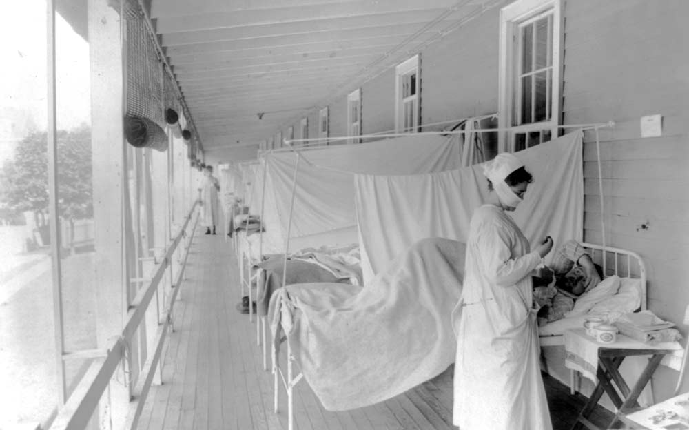 Black and white photograph of a row of hospital beds on a long open porch with sheets strung up between each bed. In the foreground, a female nurse attends to a patient in a bed.