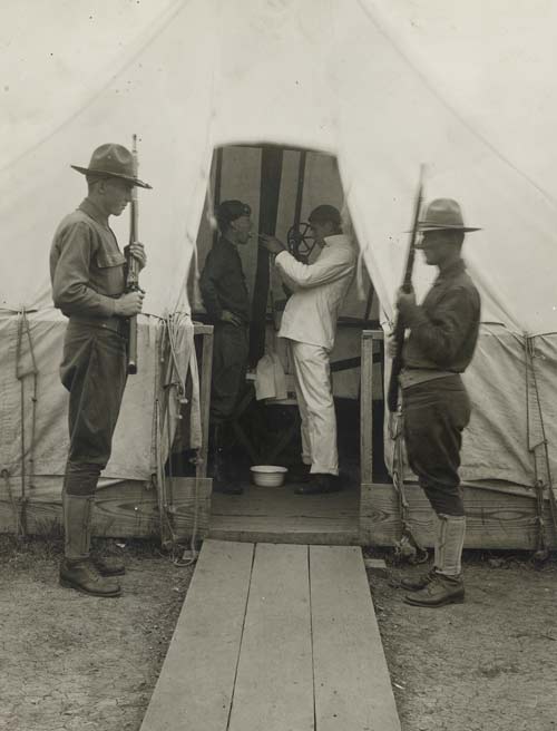 Black and white photograph of a medic tent with two soldiers standing guard outside. Inside the tent, a white-uniformed medic administers a throat spray to a third soldier.