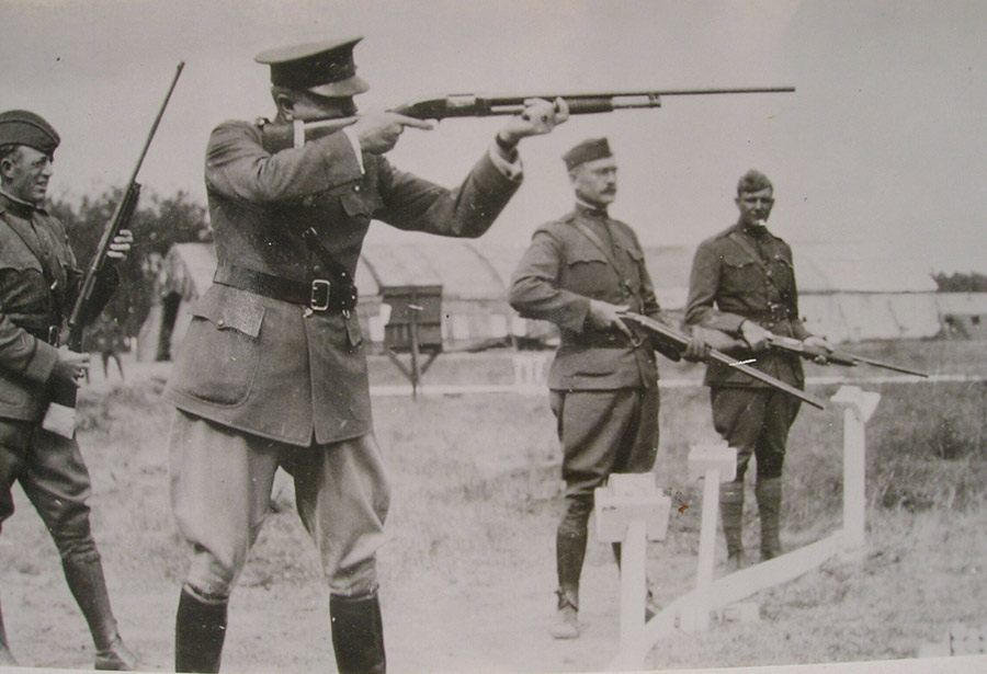 Black and white photograph of four men in military uniform holding rifles. The one in the foreground has his rifle up shooting at an unseen target. The other three are observing.