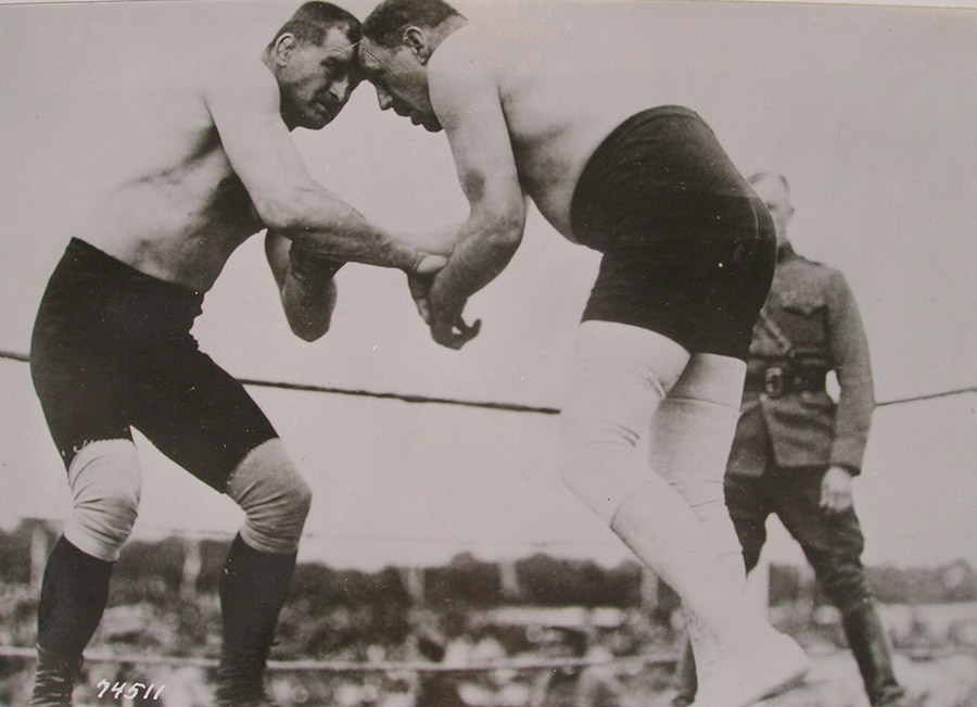 Black and white photograph. In the foreground two shirtless men grapple with each other. In the background the referee in military uniform observes.