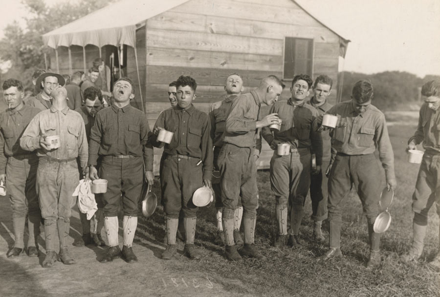 Black and white photograph of a group of men lined up with their heads back gargling salt water. They are standing in front of a shed-like structure in a field.