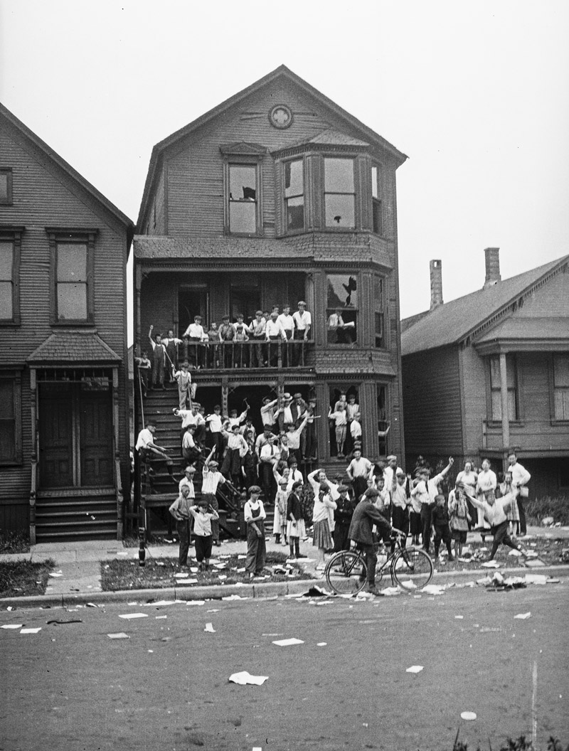 Foto hitam putih sebuah rumah di seberang jalan. Halaman depan dan terasnya penuh dengan anak-anak kulit putih. Kertas-kertas dan puing-puing berserakan di sekitar tempat kejadian.