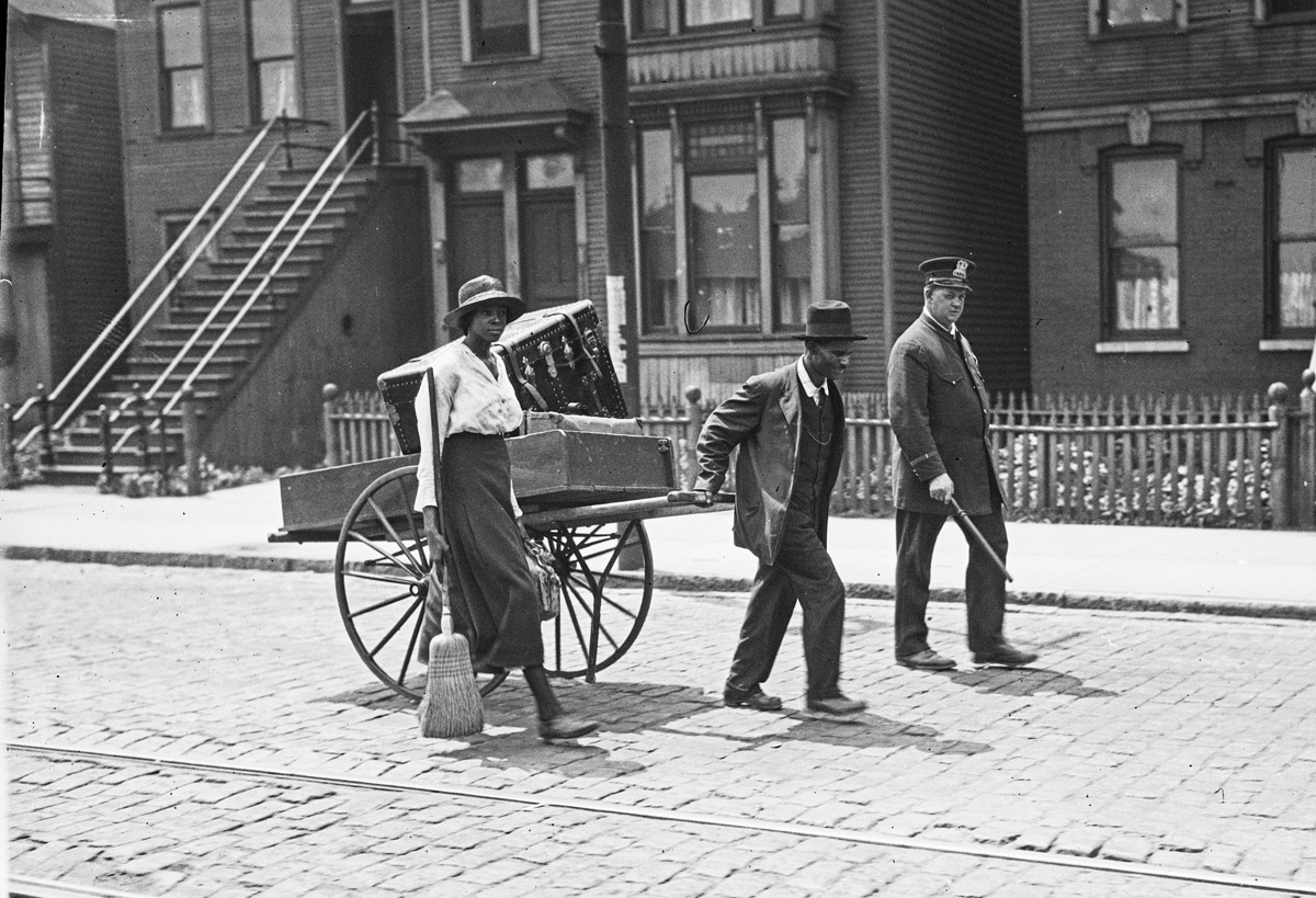 Black and white photograph of a residential street. A Black woman and a Black man walk down the street. The woman is carrying a broom and a valise. The man is pulling a cart with a large trunk and other objects in it. They are accompanied by a white man dressed in law enforcement uniform.