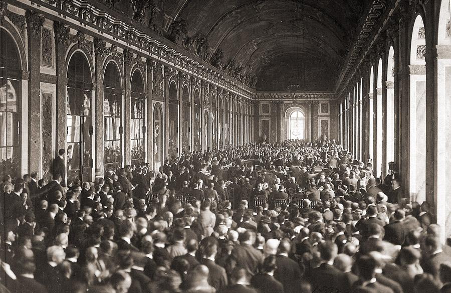 Black and white photograph of the Versailles palace Hall of Mirrors filled with a large crowd surrounding a table of seated dignitaries.