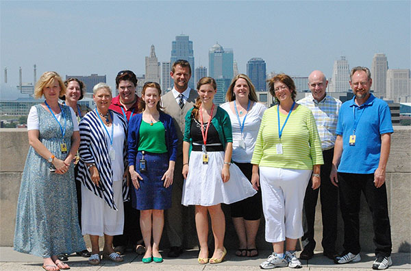 Modern photograph of a small group of adults posing for a photo on the terrace of the Liberty Memorial with the Kansas City skyline in the background.
