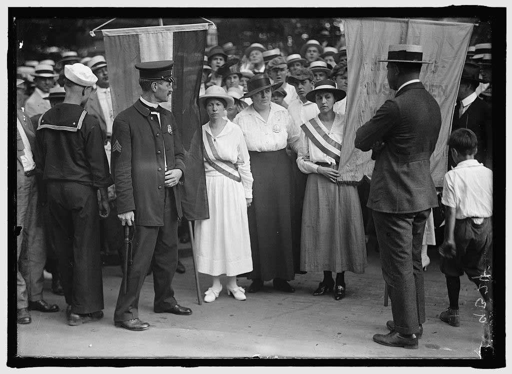 A group of women's suffrage campaigners carrying banners being blocked or confronted by uniformed men and a young boy