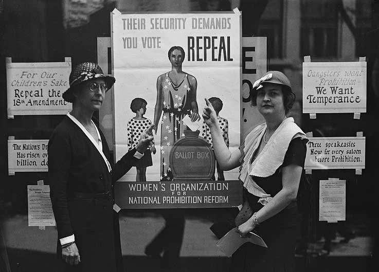 Black and white photogrpah of two women in dresses and hats pointing to a large poster that states 'Their Security Demands You Vote Repeal'