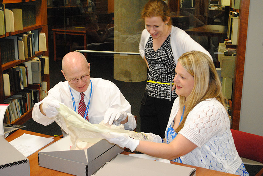 Photographie de 3 adultes blancs d'aujourd'hui vêtus de vêtements décontractés d'affaires dans une bibliothèque de recherche. Ils examinent un artefact textile.