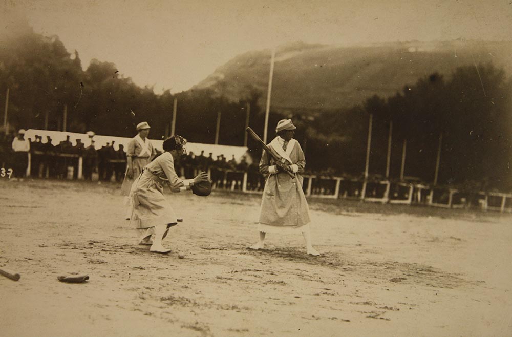Sepia photograph of a baseball player at bat with a catcher behind her. Both players wear dresses and hats.