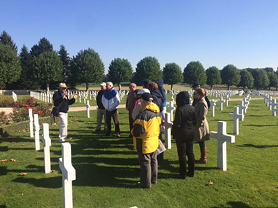 Modern photograph of a group of tourists listening to a tour guide while standing in the midst of rows of white crosses in a green lawn.