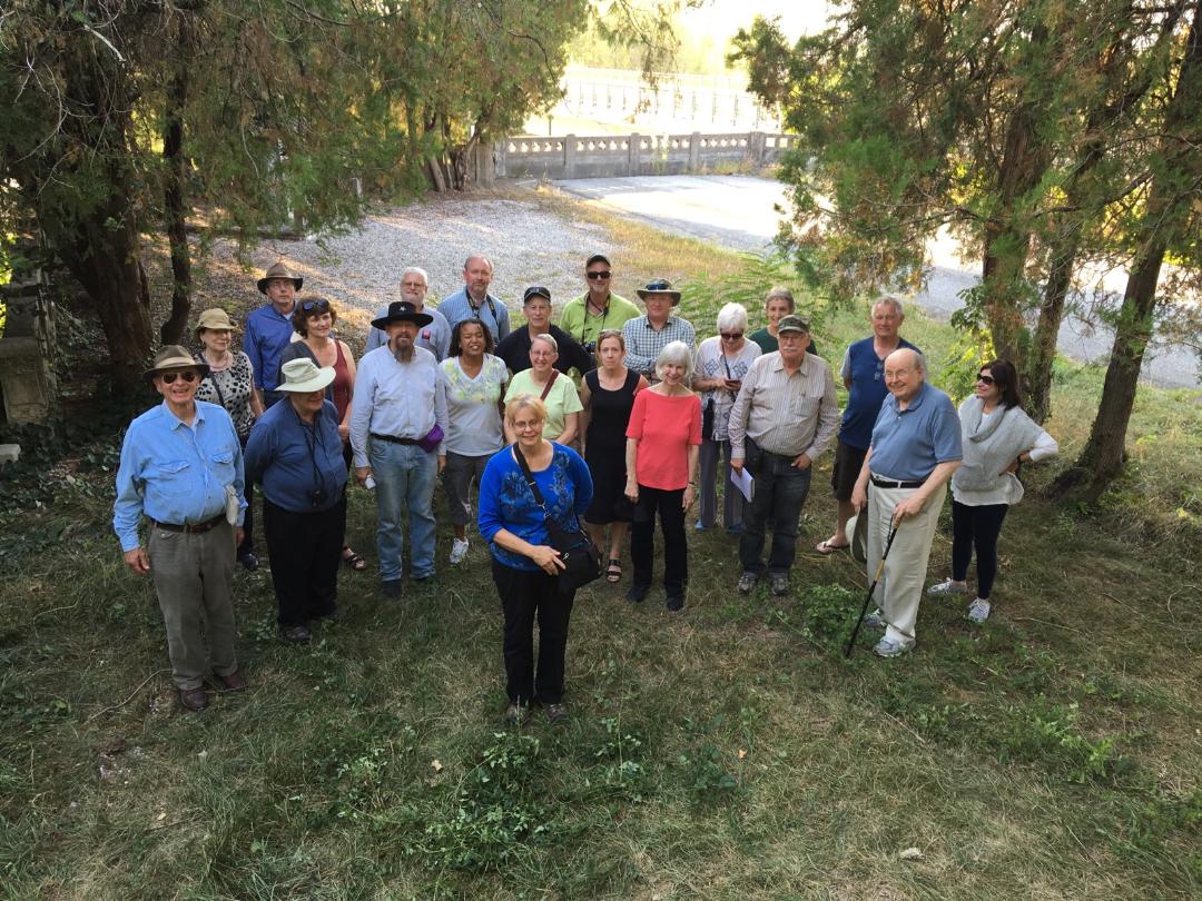 The tour group posing in front of an old bridge in a grassy wooded area.