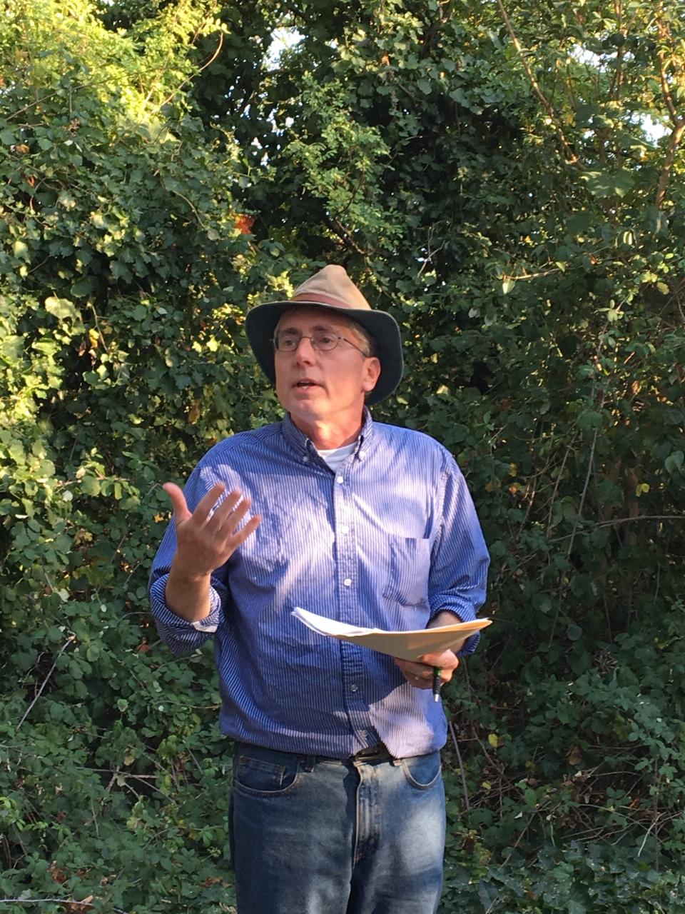 Photo of a man in a fedora hat and blue button-down shirt, standing in front of some wild greenery, reading out loud from a sheaf of papers.
