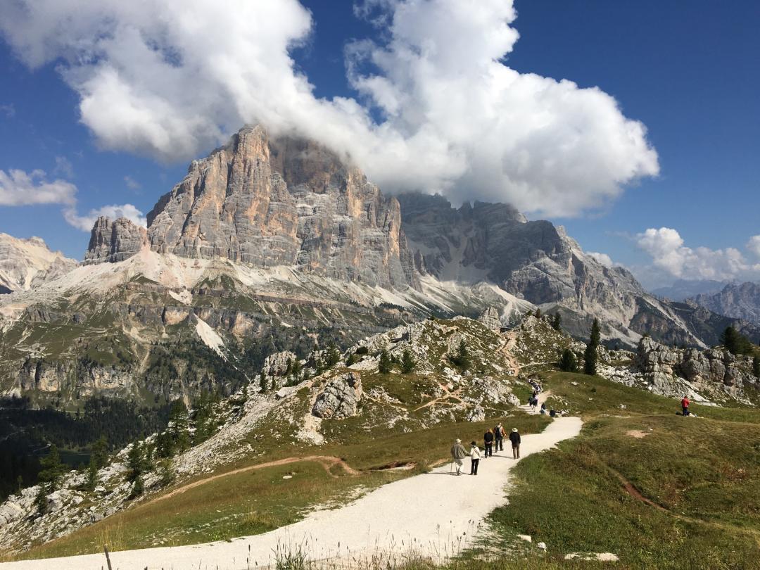 Very small humans hiking up a trail in a green valley between mountain peaks.