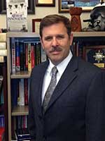 Headshot of a white man with brown hair and mustache wearing a dark grey suit jacket