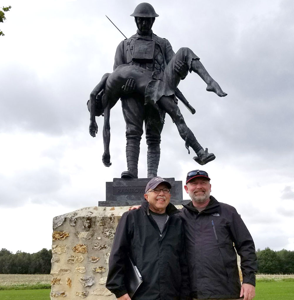 Two men posing in front of a bronze statue on a stone plinth. The statue is of a soldier carrying an unconscious person in his arms.