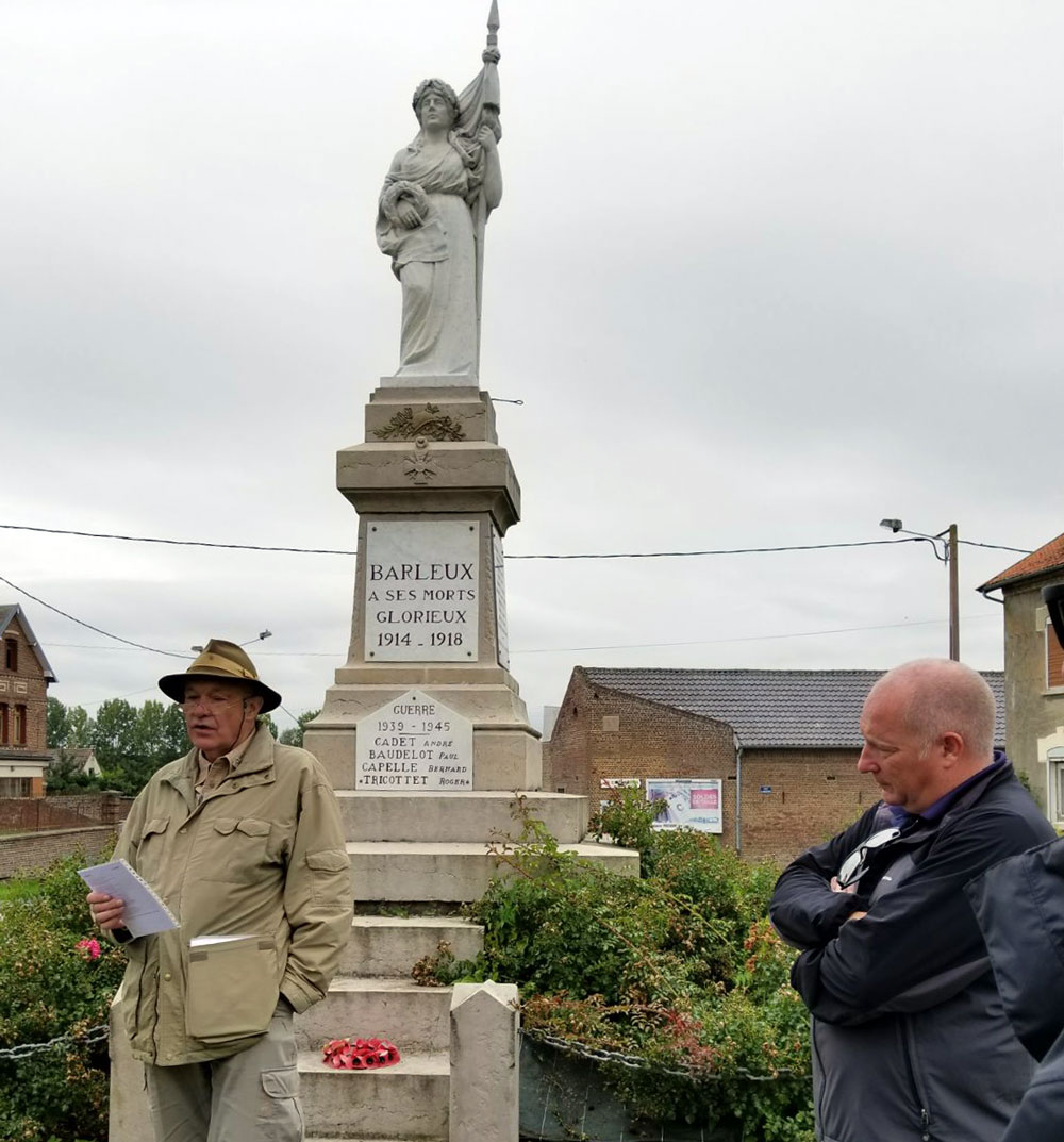 Photograph of a man standing in front of a statue of a lady dressed in classical robes. The man is reciting something from a sheet of paper.