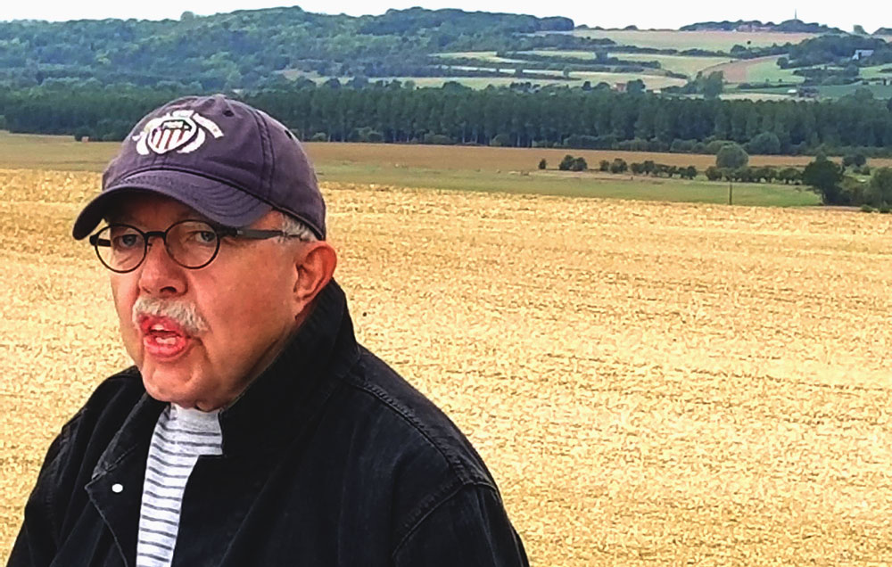 Photograph of an older white man in a baseball cap talking to someone out of frame. Behind him is a field of yellowing grass.