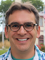 Headshot of a middle-aged white man with brown hair and glasses wearing a blue plaid button-down shirt